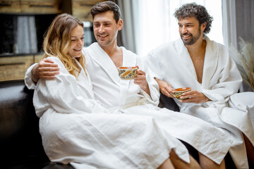 Company of friends in bathrobe sitting on sofa in living room of spa house and drink a tea. Relax and wellness concept. Spending and enjoy time together.