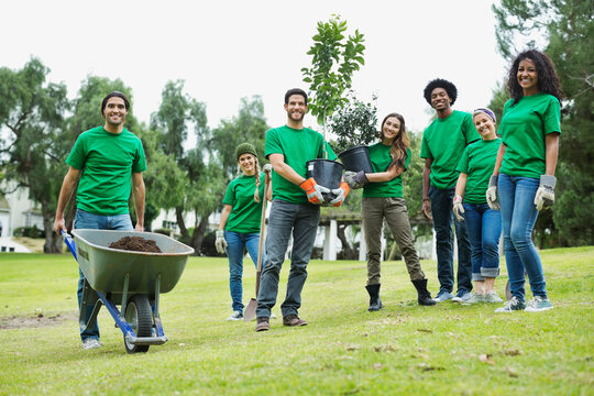 Group of happy environmentalists with potted plants and wheelbarrow in park