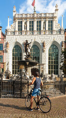 A man in shorts standing on his bike and admiring the neptun's fountain, located in the old Town of Gdansk, Poland. Tall white building in the back. Clear and bright day. Discovering the city by bike. © Chris