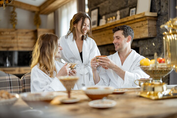 Company of adult friends in bathrobe sitting on the served table in russian style with samovar in bathhouse. Relaxing and enjoying in spa together. Wellness and leisure.