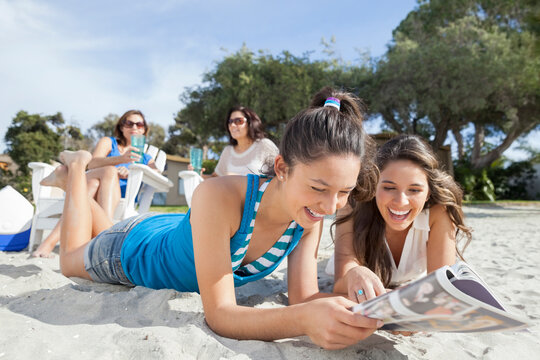 Sisters Lying On Beach Reading Magazine With Mom And Aunt In Background