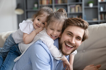 Fototapeta premium Portrait of overjoyed young Caucasian father and two small daughters hug and cuddle, relax together at home. Smiling dad have fun play with cute little girls children on family weekend together.