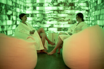 Two women in bathrobe sitting and relaxing in salt room with green light. Health and wellness spa....