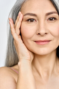 Vertical portrait of middle aged Asian woman's face with perfect skin. Older mature lady touching pampering face with hand. Advertising of cosmetology salon rejuvenating spa procedures skincare.