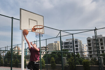 Basketball player throws basketball to a hoop. Young adult man plays basketball at outdoor court. Basketball game at public sports court