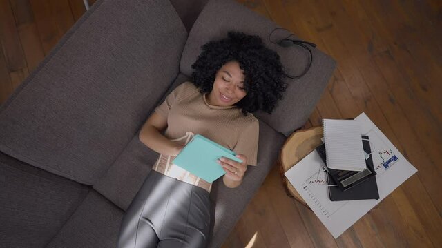 Charming satisfied African American woman lying on couch using tablet smiling. Top view of positive confident young beautiful manager working online surfing Internet in home office living room