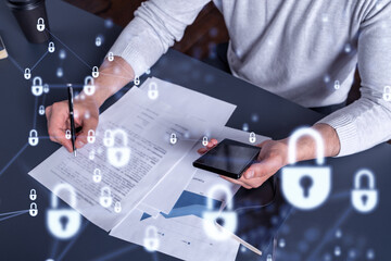 A businessman in casual wear signing the contract to prevent probability of risks in cyber security. Checking the phone. Padlock Hologram icons over the working desk.