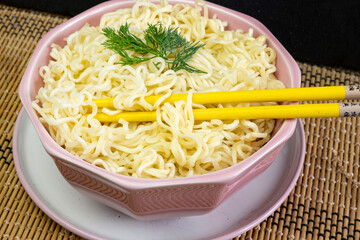 Quick instant noodles with parsley in a pink bowl on  grey tablecloth, Asian Chinese ramen pasta fast food eaten with yellow bamboo chopsticks