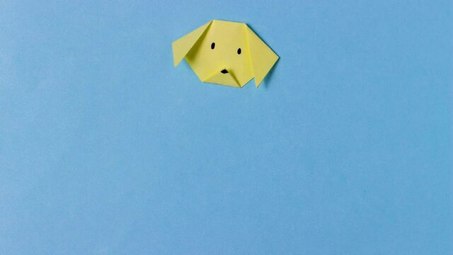 The dog's head is folded from yellow paper in the technique of origami with painted mustache. In center of the blue background. Concept of love dogs, pets, pastime, hobbies, activities with children.