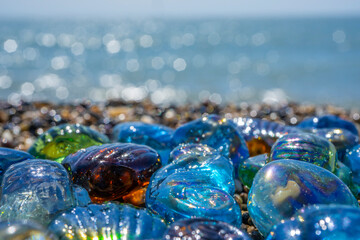 Beautiful shape glass and stones on the seashore. Azure clear sea water with waves. Green, blue shiny glass with multi-colored sea pebbles close-up. Beach summer background. High quality photo