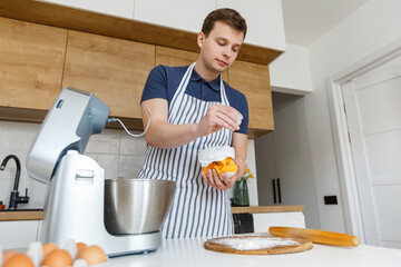 Fototapeta na wymiar Young handsome man in apron sifting flour in modern kitchen. Concept of homemade bakery food, male cooking and domestic lifestyle. Chef making pastry using food processor at home
