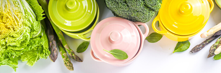 Various organic green spring vegetable ingredients with empty colorful portioned cooking pots on white kitchen table, top view. Diet healthy soup, stew food preparation concept