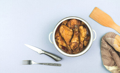 Cuisine workspace concept with saucepan filled with roasted fried chicken, top view of table with towel and wooden spoon
