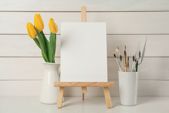 Easel with blank canvas, brushes and tulips on white wooden table