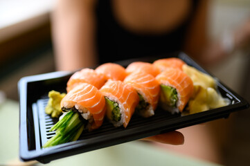Womans hand holding fresh takeaway philadelphia sushi rolls closeup with distinct salmon texture fresh rise avokado and cucumber served in black plastiv box