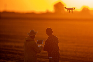 Fototapeta premium Two farmers using drone surveying field at sunset