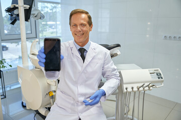 Friendly dentist holding a smartphone with blank screen