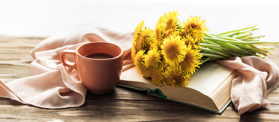 The atmosphere of a romantic morning, coffee in bed. A bouquet of yellow dandelions on an open book and a cup of coffee on a pink silk bed. Home interior. Lifestyle,selective focus.	