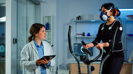 Woman with mask running on cross trainer testing heart rate using electrodes, medical researcher...