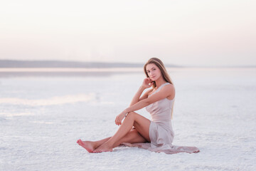 Fototapeta na wymiar Young blonde woman in an evening airy pastel pink powdery dress sitting barefoot on white crystallized salt. Girl with natural make-up, hair is developing. Salt mining trip, walking on water at sunset