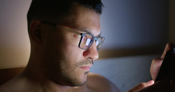 Young man in bed looks at the screen of his smartphone. The face is illuminated by a bright screen, reflected in the glasses.