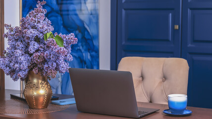 A cup of tea or coffee on a working wooden table, a laptop, a phone, a bouquet of lilacs in a gold vase. Cozy interior with a blue accent. Working office desk, working at home. 