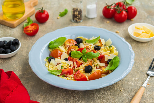 Salad with orzo pasta, tomatoes, olives and parmesan in a gray plate on a gray concrete background. Pasta salad recipes.