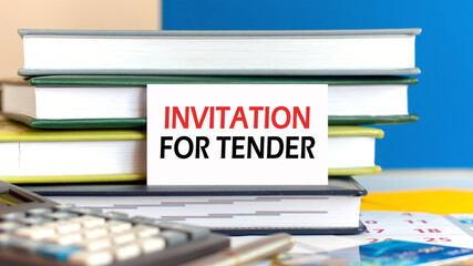 white card with the text INVITATION FOR TENDER stands against the background of books stacked blue background, defocus