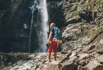 Man with backpack dressed in active trekking clothes holding trekking boots in hand near mountain...