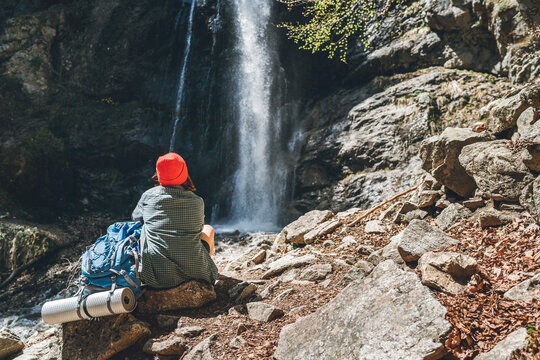 Woman with a backpack in red hat dressed in active trekking clothes sitting near the mountain river waterfall and enjoying the splashing Nature power. Traveling, trekking and nature concept image.