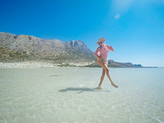 Cheerful woman enjoying jumping and splashing in shallow water in sea. She is in turquoise water by...
