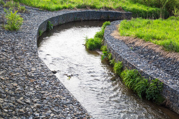 A channel made of granite stones for the river stream with grass and shrubs on the banks. Landscaping of the city, river hydrology. - obrazy, fototapety, plakaty