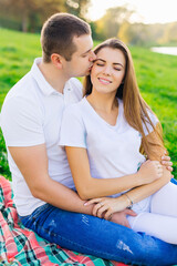 In the park, on the green grass, a cute affectionate couple is sitting, a man gently kisses a woman.