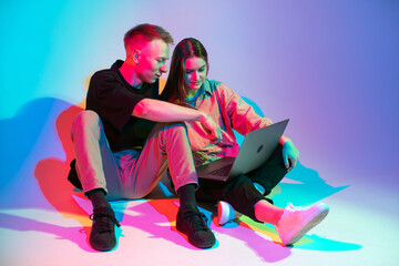 Stylish young couple with laptop working, watching series or shopping online. Colorful neon light