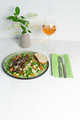 salad with fried beef, vegetables, sauce on green plate on white background, rose wine glass, bird cherry blooming branch, fork and knife on textile