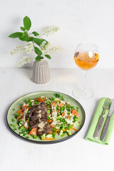 salad with fried beef, vegetables, sauce on green plate on white background, rose wine glass, bird cherry blooming branch, fork and knife on textile