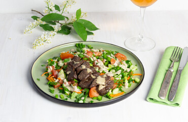 salad with fried beef, vegetables, sauce on green plate on white background, rose wine glass, bird cherry blooming branch, fork and knife on textile