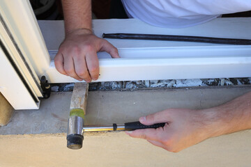 Worker installing new plastic window, using small hammer and wooden wedge