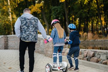 A father helps and teaches his young children to ride a Segway in the Park during sunset. Family vacation in the Park.