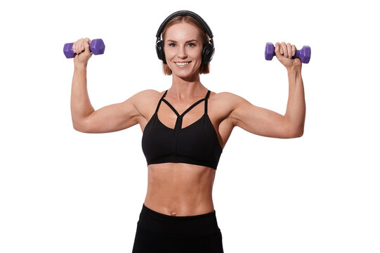 Strong muscular sportswoman in headphones and black outfit doing exercise with dumbbells over white isolated background © khmelev
