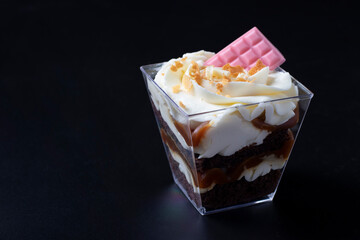 Trifle made from chocolate biscuit with caramel-peanut filling on black background. Dessert is decorated with creamy cheese cream and pink chocolate.