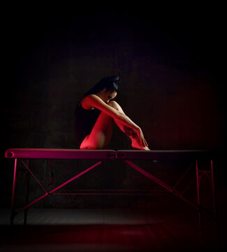 Young Sexy Brunette Woman With Tattoo On Buttocks In Bodysuit Sits On Massage Table Hugging Her Knees, Looking Down Over Dark Background