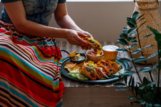 Latin American Woman In Traditional Colorful Skirt Eating Chicken Tacos, Nachos, Salsa And Guacamole