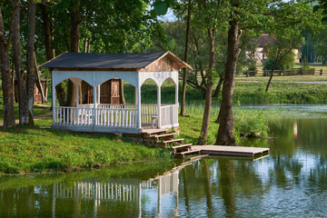 Obraz premium Summer gazebo on shore of pond. Wooden pergola in Merechevshchina village, Tadeusz Kosciuszko birthplace, Brest region, Belarus.