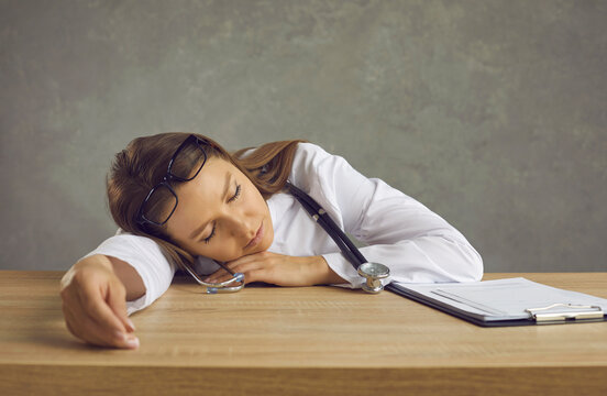 Tired Young Woman In White Lab Coat With Stethoscope Sleeping Sitting At Uncomfortable Work Desk In Office Room. Exhausted Doctor, Nurse Or Emergency Medical Technician Resting After Long Night Shift