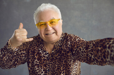 Funny rich senior man in bling gold bracelet and trendy glasses saying Call Me. Goofy old guy in glamorous leopard patterned shirt and cool yellow sunglasses taking selfie and doing phone call gesture