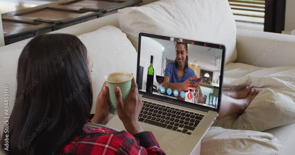 Sticker African american woman holding a coffee cup having a video call on laptop sitting on couch at home