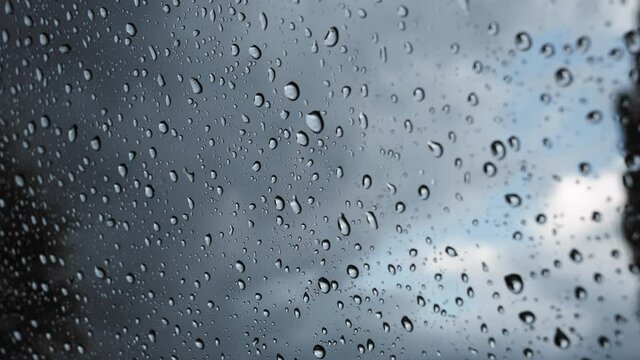 Water raindrop on a clear window glass. Raining on a car windscreen on a moody day. Moving droplets close up in front of a dark cloudy sky. Liquid fluid splashing on the windshield.