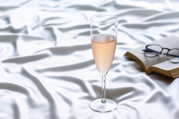 A glass of Rose wine on wavy satin cloth with book and spectacles. Stay home concept.