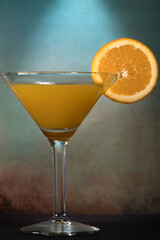 Martini glass with a mix of orange juice and alcohol with a slice of orange fruit against dark and moody orange blue background